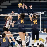 Two T-Birds go up for the block, wearing blue jerseys, while a Saint Mary's player attempts to clear them, wearing a white jersey.