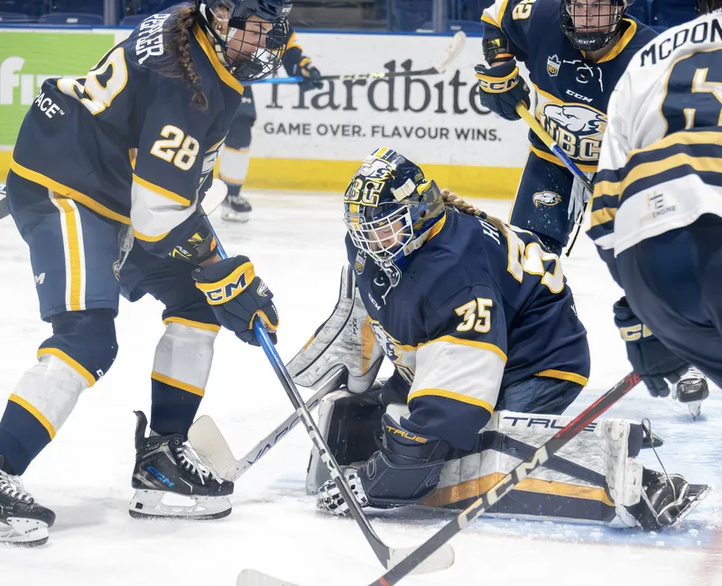T-Birds goalie Elise Hugens controls the puck in front of the net, wearing a blue jersey, facing off against the Spartans in white.
