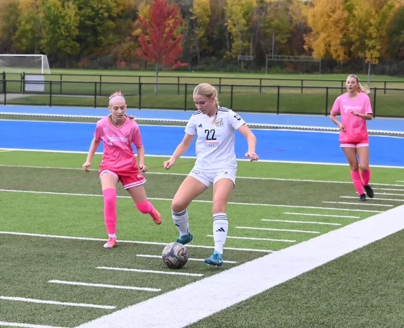 A UBC player controls the ball with two defenders in pink jerseys nearby.