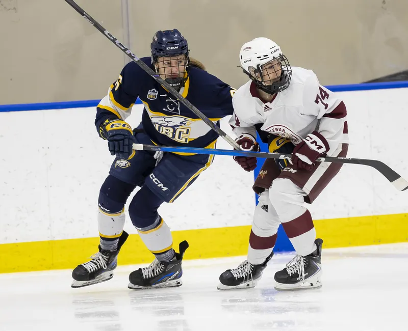 A Thunderbird in a blue and white jersey tries to skate past a MacEwan player in white and red.