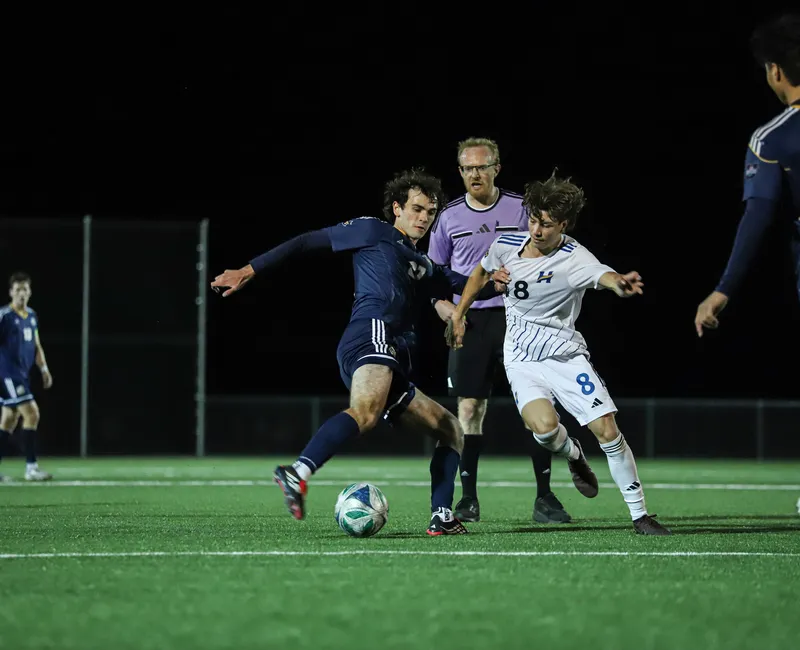 A UBC and UBCO player grapple for possession of the ball as the referee looks over.