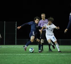 A UBC and UBCO player grapple for possession of the ball as the referee looks over.
