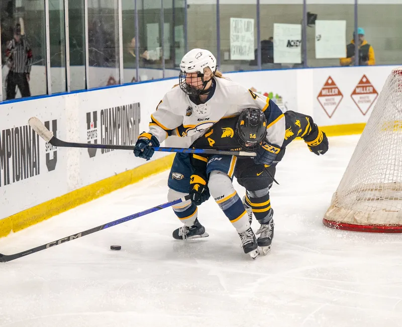 A T-Bird in a white jersey battles for the puck with a Warrior in a black jersey behind the net.