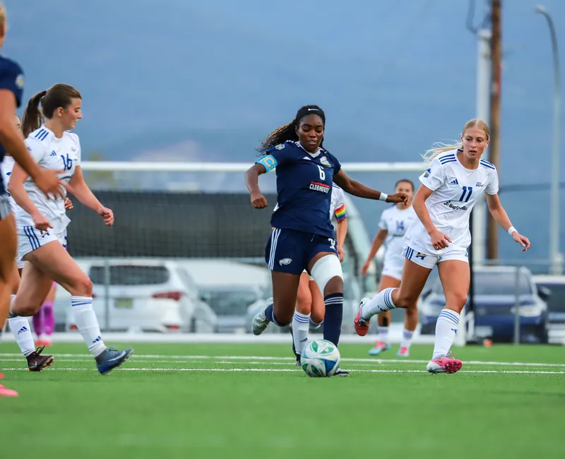 UBC's Taiya Dennehy runs up field with the ball, wearing a blue jersey. She's chased by multiple Heat players in white.
