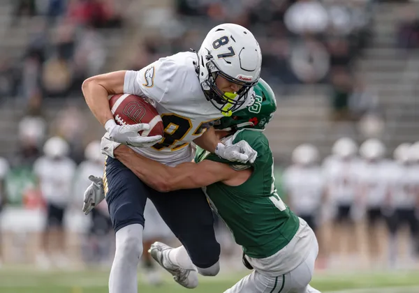 A T-Bird in white holding on to the football is tackled by a Huskie in dark green.