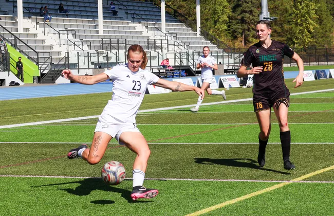 Norman, in a white T-Birds jersey, takes a shot with a defender in a brown jersey behind her.