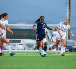 UBC's Taiya Dennehy runs up field with the ball, wearing a blue jersey. She's chased by multiple Heat players in white.