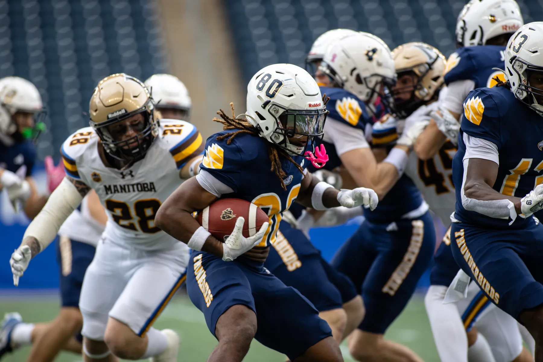 Surrounded by both Manitoba players in white jerseys and UBC players in blue jerseys, a T-Bird runs to the right, with the ball in his hand.
