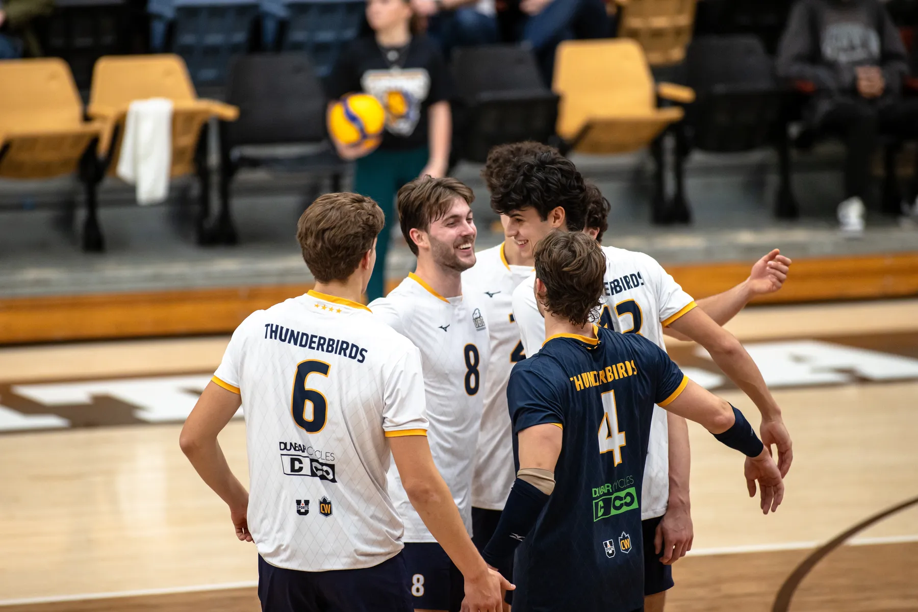 On an indoor volleyball court, a group of 5 T-Birds celebrate in a group hug, with most players in white, and one player in dark blue.