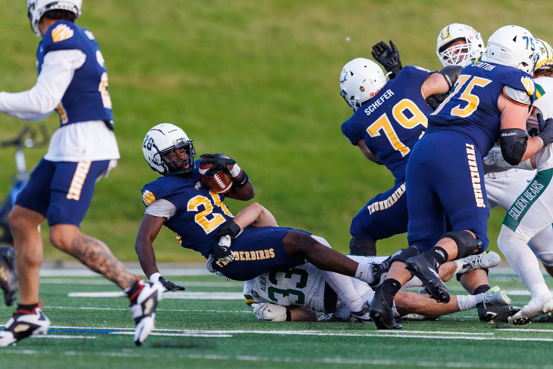 A UBC player in blue is tackled by an Alberta player in white, with numerous other UBC players around blocking other Alberta players.