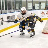 A T-Bird in a white jersey battles for the puck with a Warrior in a black jersey behind the net.