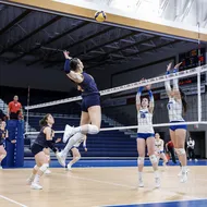 A UBC player in blue jumps up to spike the ball, while two Montreal players in white go up at the end to match her.