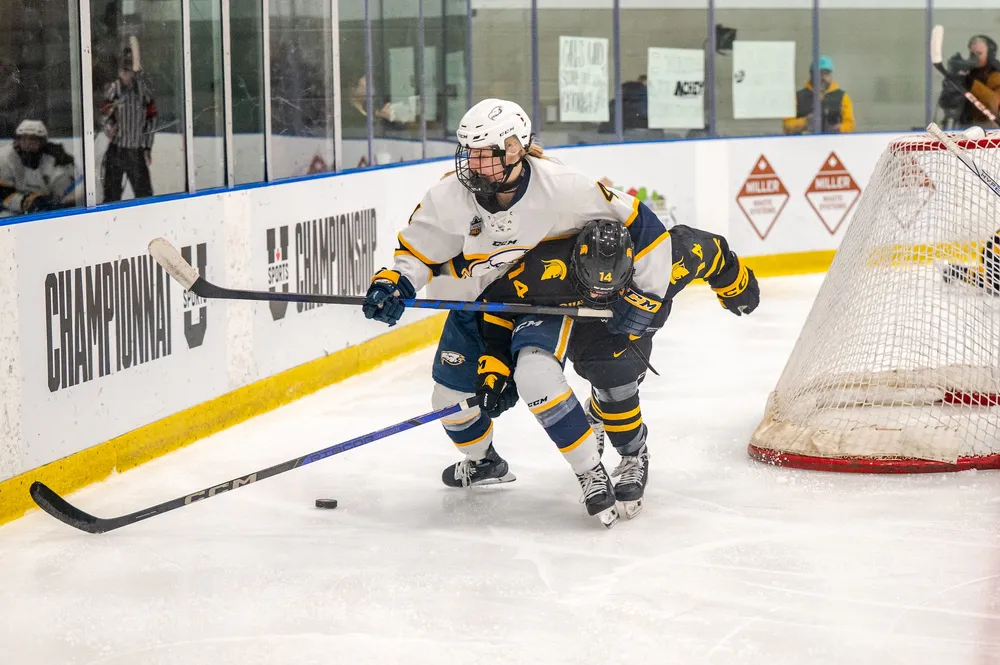 A T-Bird in a white jersey battles for the puck with a Warrior in a black jersey behind the net.