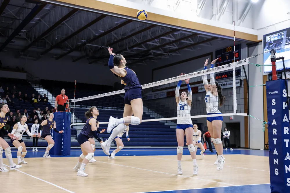 A UBC player in blue jumps up to spike the ball, while two Montreal players in white go up at the end to match her.