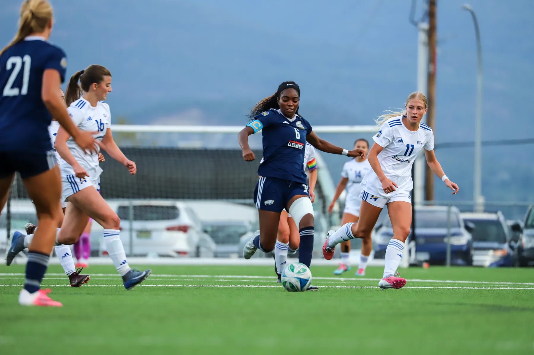 UBC's Taiya Dennehy runs up field with the ball, wearing a blue jersey. She's chased by multiple Heat players in white.