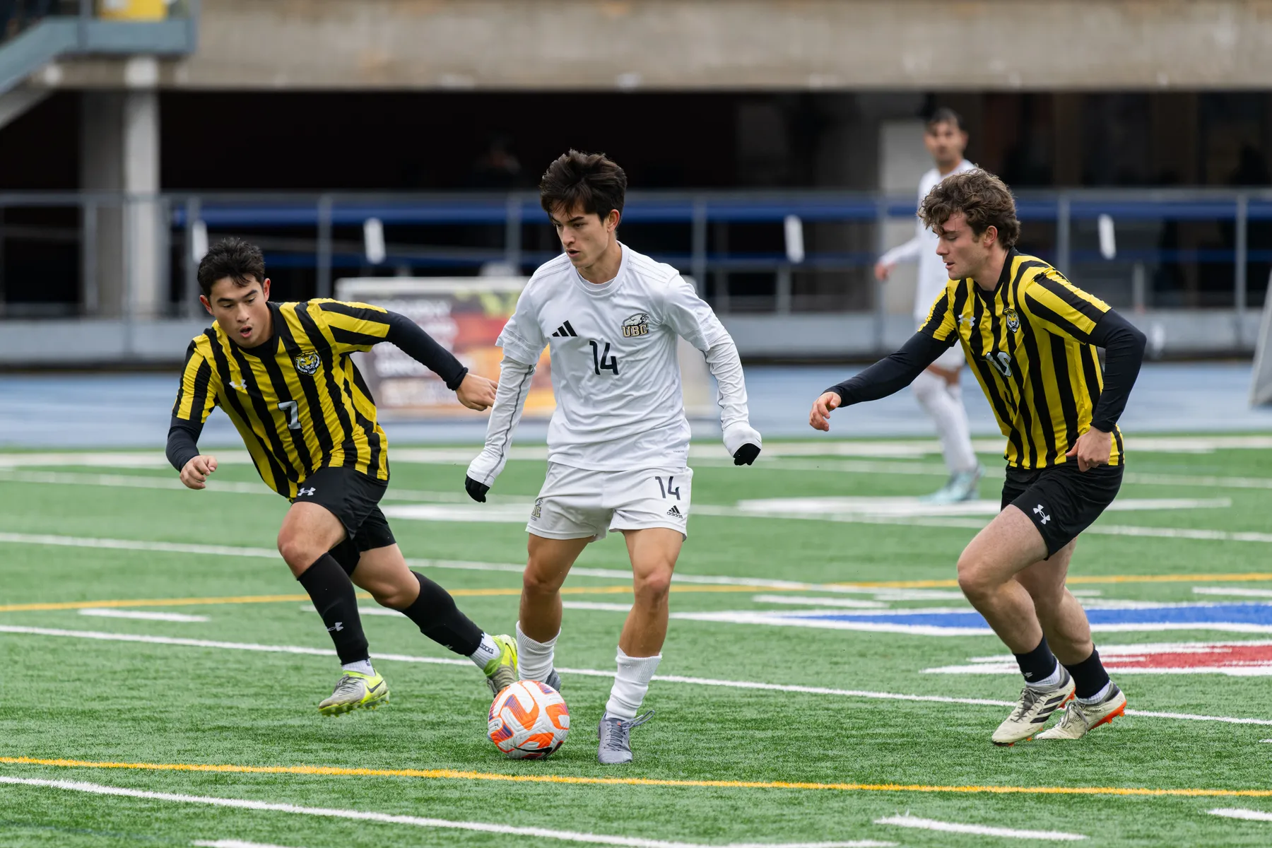 A T-Bird in a white jersey runs with the ball, with Dalhousie players in black and yellow vertical striped jerseys on both sides of him.