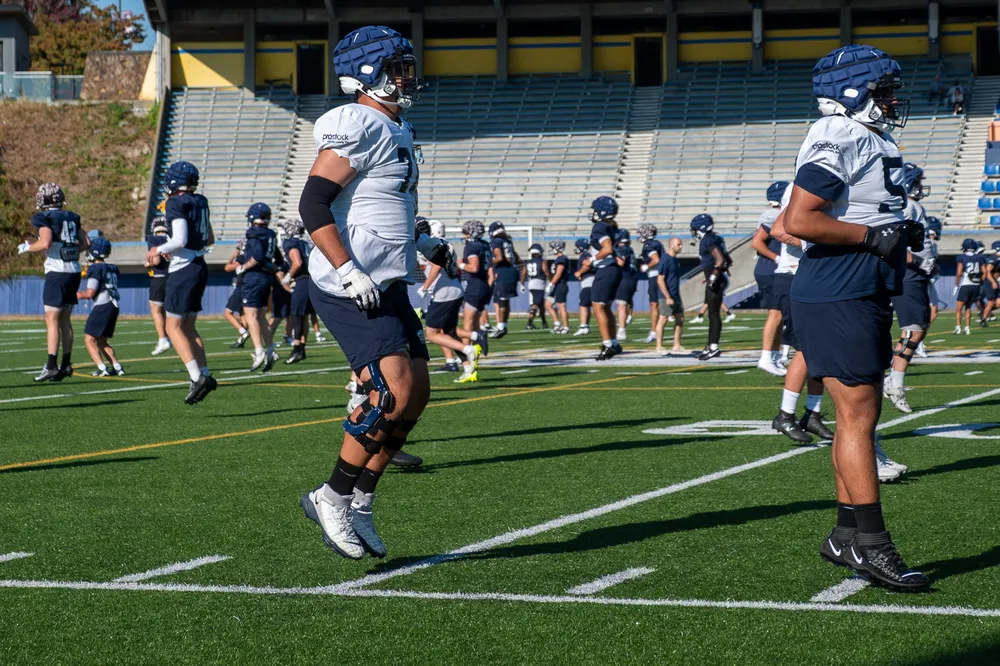 Two linemen walk in front of the camera at practice, with the rest of the team visible behind them.