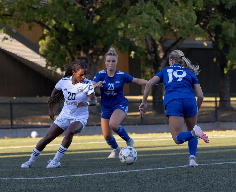 Thompson attempts to control the ball, while wearing a white jersey, while two UBCO players in blue attack the ball.