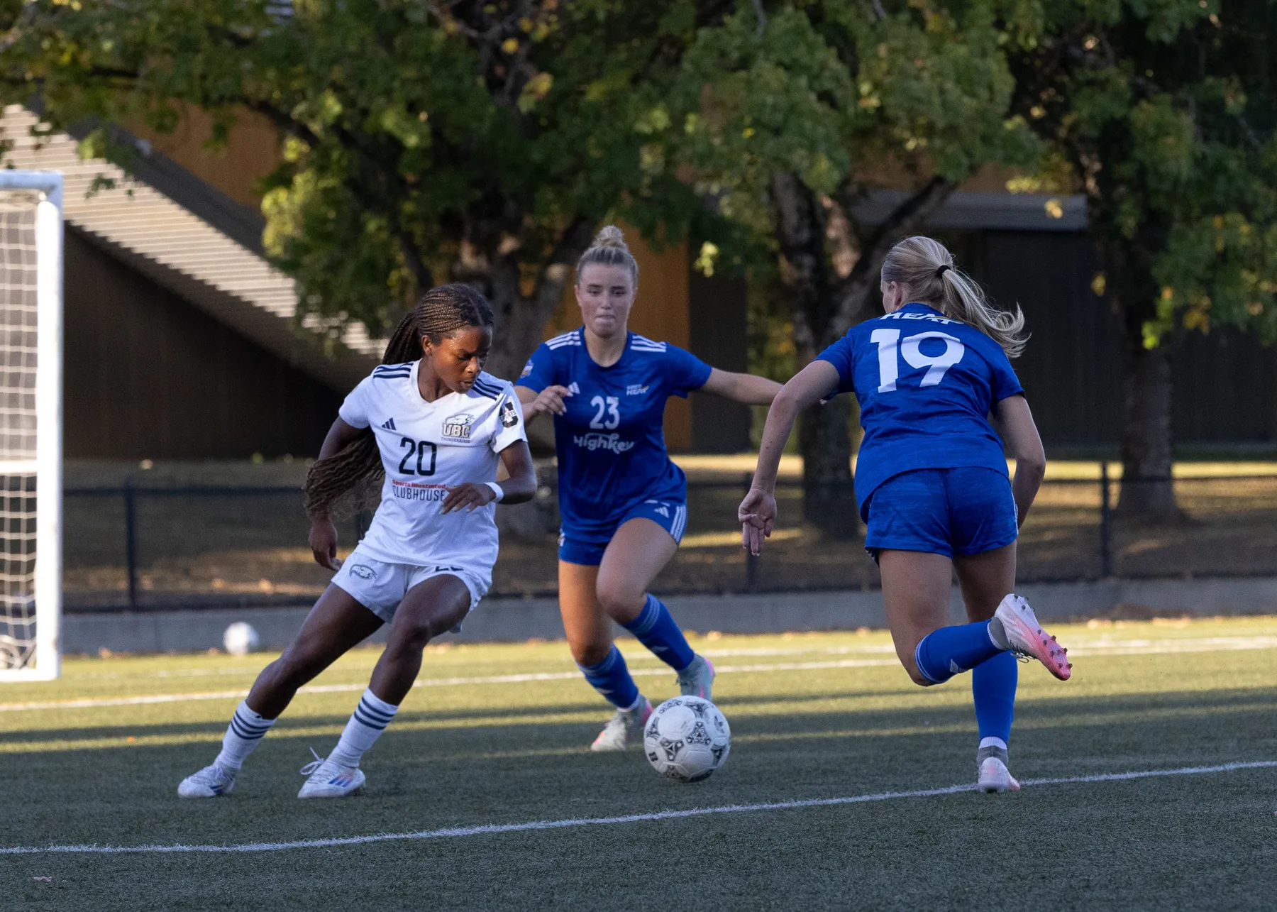 Thompson attempts to control the ball, while wearing a white jersey, while two UBCO players in blue attack the ball.