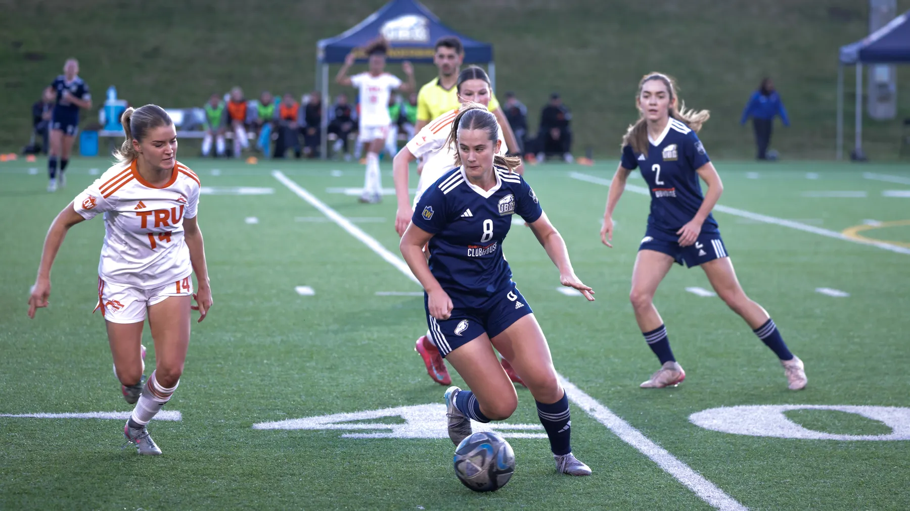 A UBC player in blue runs away from two TRU players in white, while dribbling the ball.