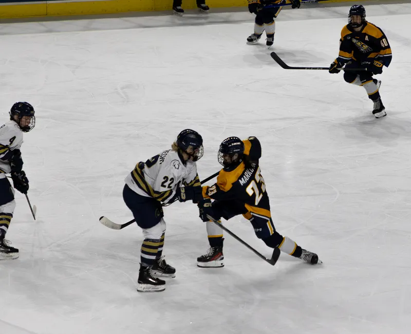 A Thunderbird in an alternate blue and gold jersey shoots the puck with a Trinity Western defender, in white, in her face.