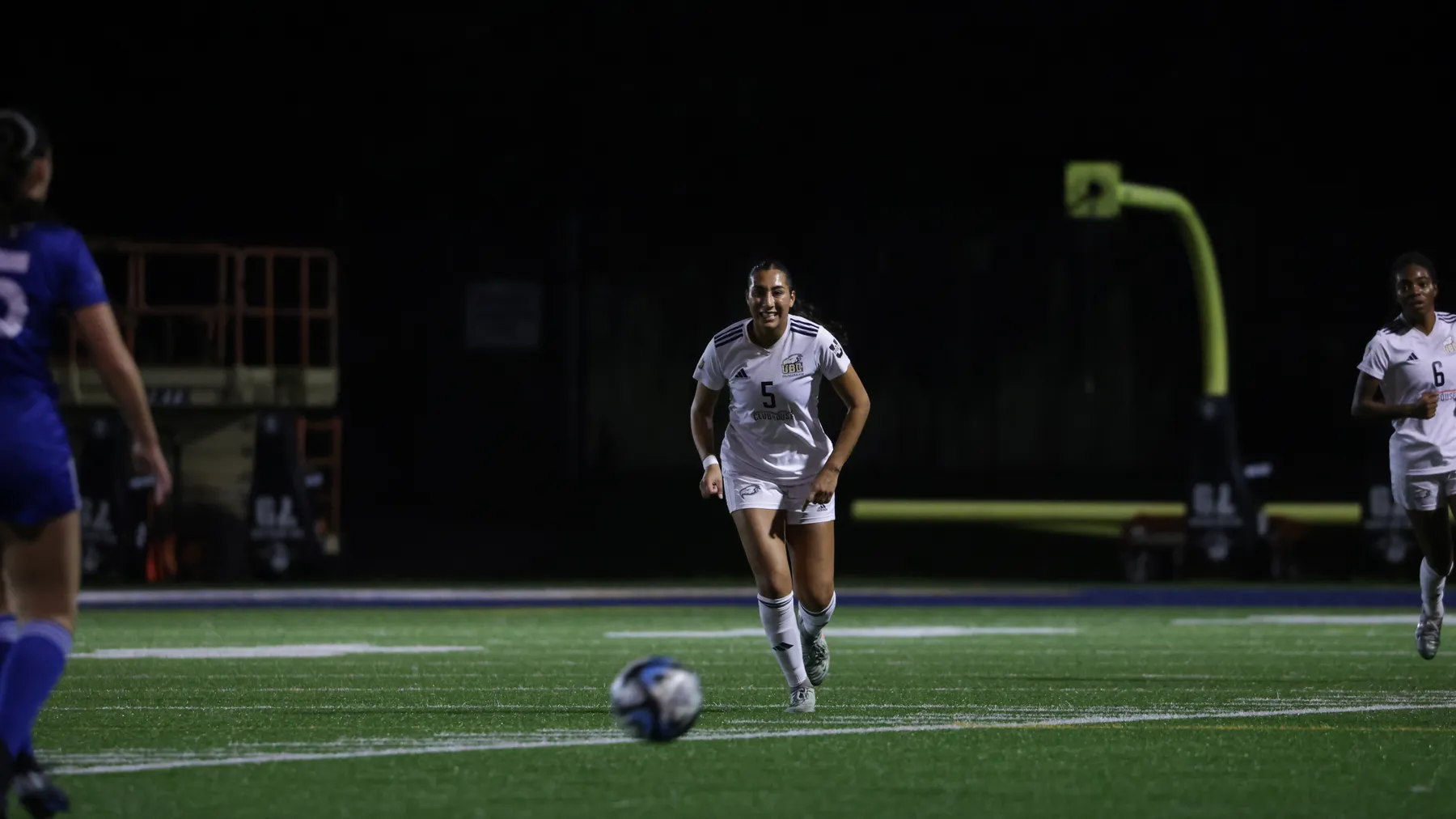 At night, two UBC players in white chase down the ball, which is rolling down the field, towards the camera.