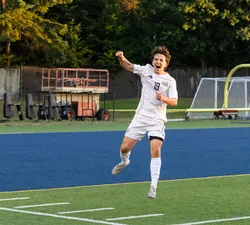 A UBC player in a white jersey jumps up in the air, pumping his fist in celebration.