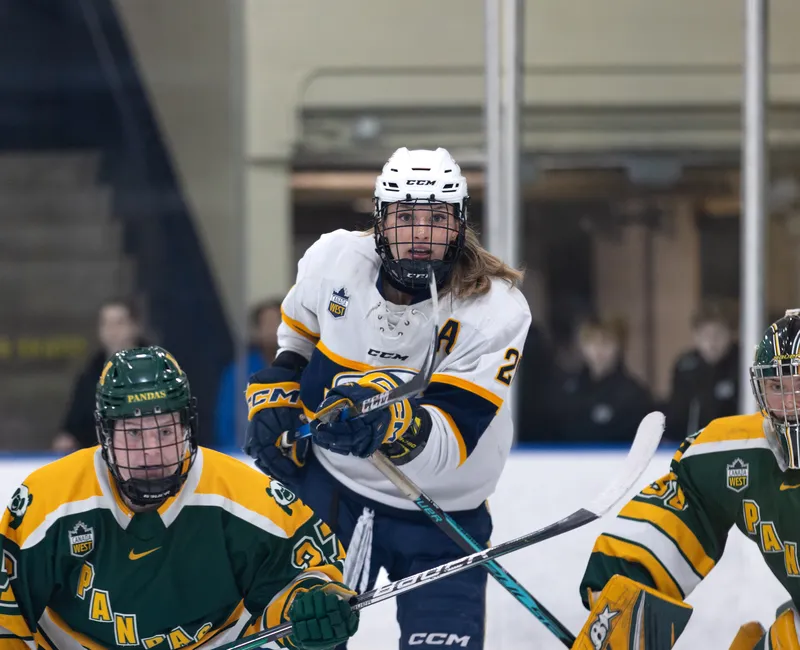 Elliott — in a blue and white jersey — finds space in front of the net of the Alberta Pandas, with a Pandas defender and goalie nearby in green and yellow.