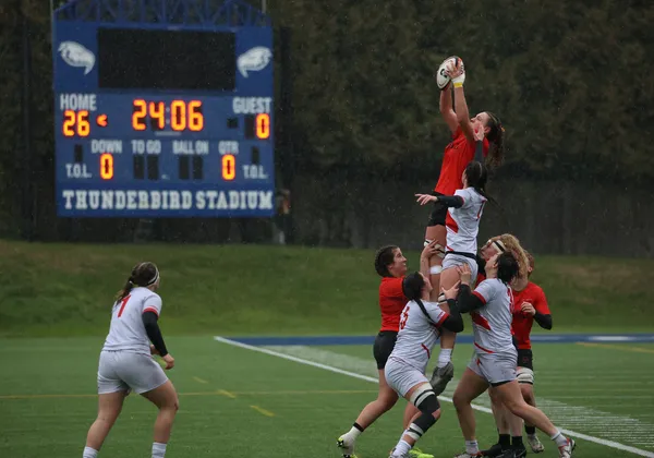 With the scoreboard illuminated behind them, a Laval player in red is lifted by her teammates to catch the ball, while surrounded by Acadia players in white