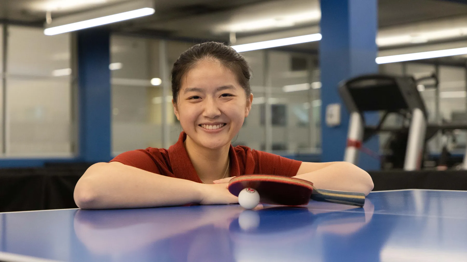 Ivy Liao sits by a table tennis table, holding a paddle and a ball.