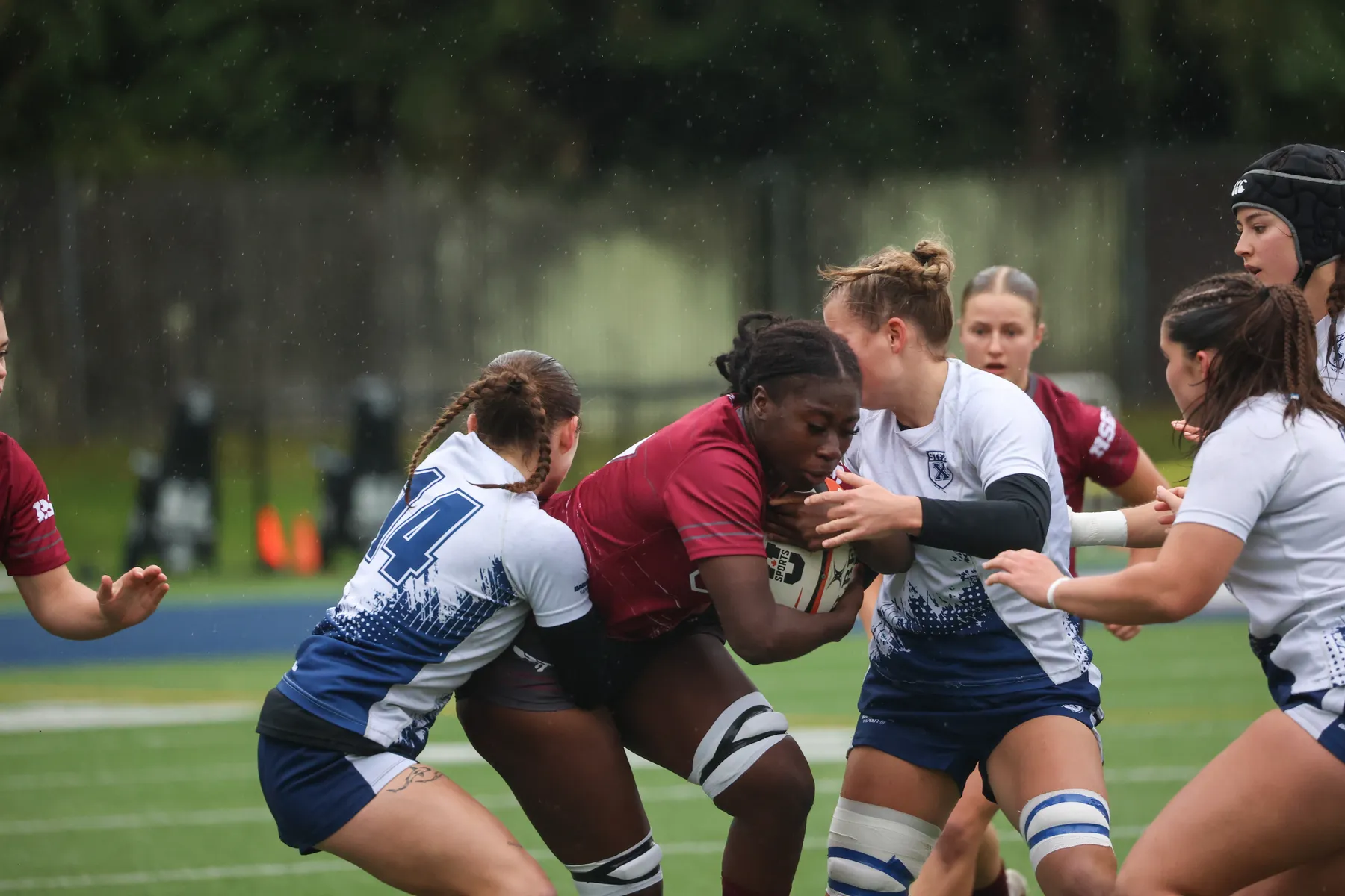 An Ottawa player in burgundy, holding the ball, is tackled by two STFX players in white and blue.