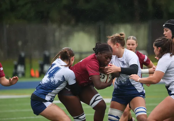 An Ottawa player in burgundy, holding the ball, is tackled by two STFX players in white and blue.