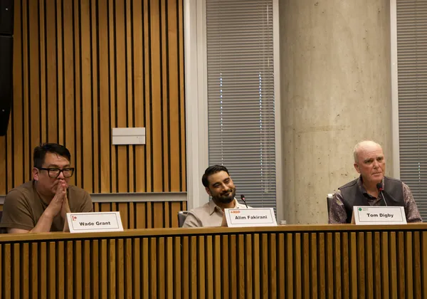 A photo of three men sat at a desk with mics in front of them and white placards of their name.