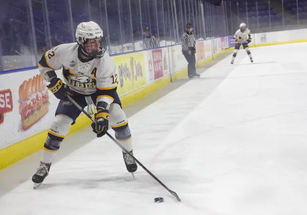 A hockey player in a white jersey kit skates with the puck near the boards of an indoor rink.