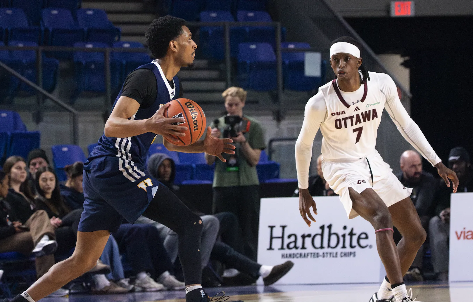A male basketball player in a navy blue jersey dribbles while a player in a white jersey titled "Ottawa" defends.
