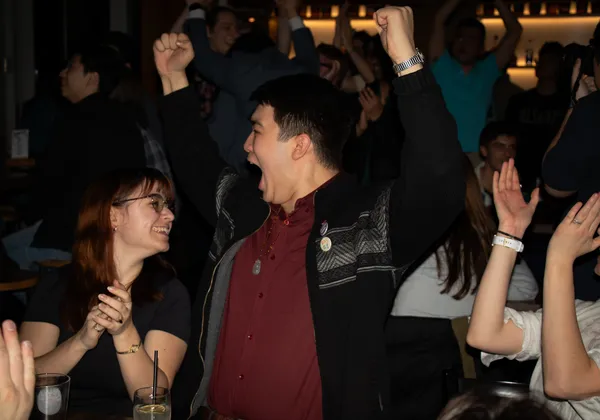 Yi-Kieran cheers with their arms up as people around them clap and smile.
