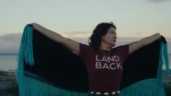 A person in a land back shirt with a cape spins against a setting sun
