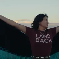 A person in a land back shirt with a cape spins against a setting sun