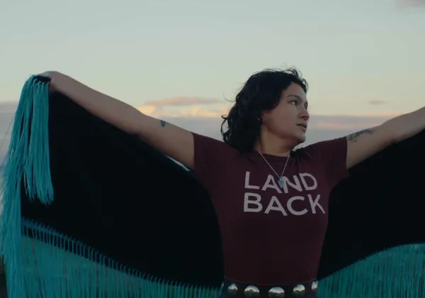 A person in a land back shirt with a cape spins against a setting sun