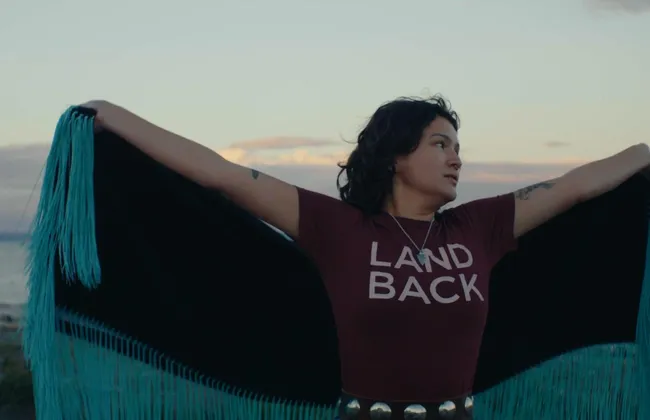 A person in a land back shirt with a cape spins against a setting sun