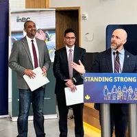 Three men stand together as one of them — UBC President Bacon — speaks and gesticulates to an audience.