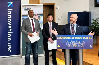 Three men stand together as one of them — UBC President Bacon — speaks and gesticulates to an audience.
