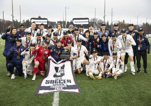 The UBC men's soccer team is huddled around a black and white banner that says "U Sports men's soccer championship 2024" in English and French. The players are in white jersey kits and the goalkeepers are in red. Support staff are wearing navy blue jackets and every individual has a gold medal around their neck and a black championship hat.