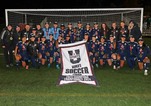 The UBC women's soccer team, clad in navy blue jersey kits, stand in two lines in front of the goalie net on an outdoor soccer field. They are holding a black and white banner that says "U Sports women's soccer 2024 Champions" in English and French and have gold medals around each of their necks.
