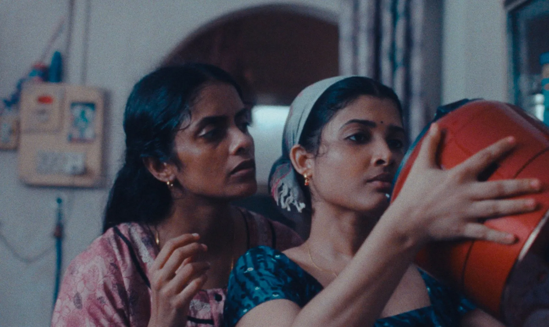 A woman in a blue top and a bandana holds a red pot while a woman in pink is behind her, watching.