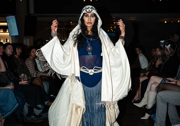 A woman walking down a runway in a dark blue translucent shirt and lighter blue skirt. Her head and arms are covered by a white shawl, and her hands are raised to eye level.