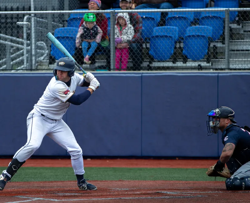 A baseball player in a white jersey kit winds up to hit the ball at home base.
