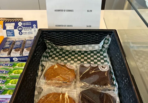 A basket of cookies individually wrapped in plastic with a sign reading "gluten-free" and a list of baked goods and their prices.