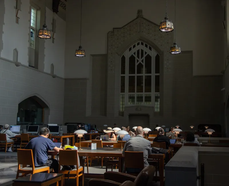 A photo of students studying in the Irving K. Barber Learning Centre, taken from the street level, who are facing away from the camera and seated a few meters away at the tables in the centre of the image.