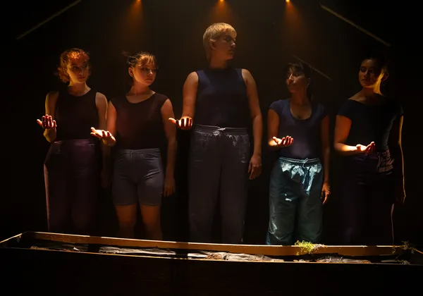 Five femme-presenting people stand in a line in a dimly lit room, each of their right arms outstretched in front of them.